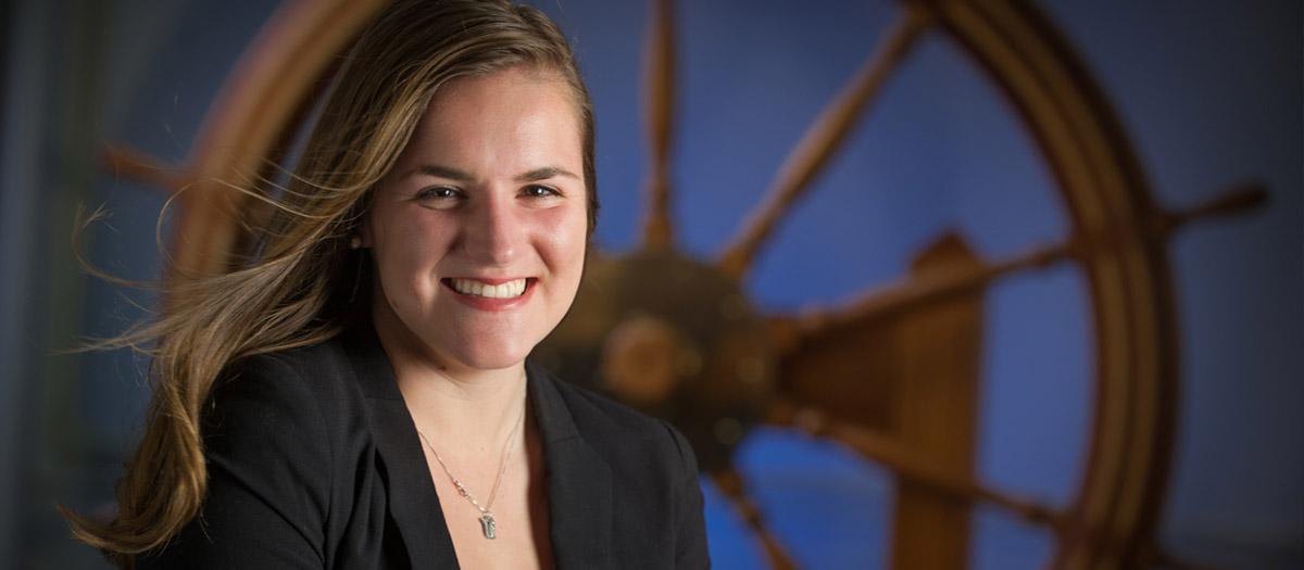 A student poses in front of a ship's steering wheel.